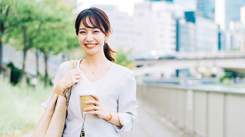 コーヒーのカップを片手に屋外を歩く笑顔の女性